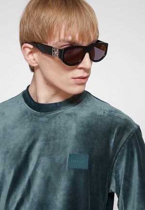 Giovane persona con occhiali da sole neri firmati e una camicia di velluto verde acqua a manica lunga con un piccolo logo "Hugo" sul petto.