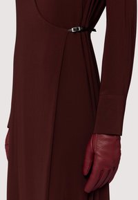 Bordowa sukienka o gładkiej fakturze, z metalowym akcentem w kolorze srebrnym w talii. Czerwona skórzana rękawiczka uzupełnia zestaw.
