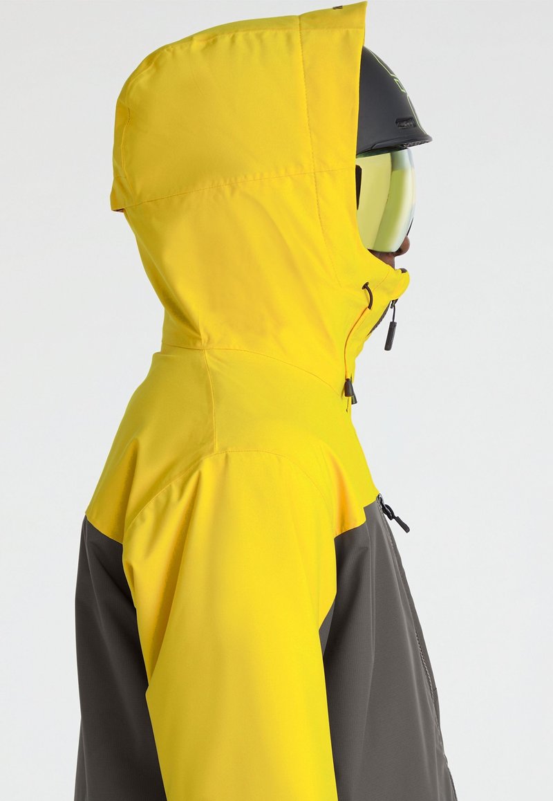 Kurtka narciarska z żółtym kapturem i górną częścią, ciemnoszarym dolnym fragmentem, z zamkami błyskawicznymi i smukłym wzornictwem.