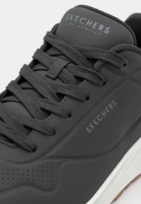 Czarne sneakersy Skechers z teksturowanym syntetycznym wykończeniem, płaskimi sznurowadłami i białą gumową podeszwą. Logo 'SKECHERS' po bokach w jasnoszarym kolorze.
