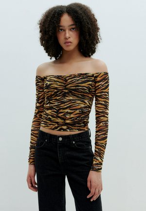 Een off-the-shoulder top met lange mouwen in een zwart en oranje tijgerprint, gemaakt van rekbaar materiaal; gecombineerd met hoge, donkere jeans.