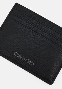 Cardholder din piele neagră, cu o suprafață texturată, multiple sloturi pentru carduri și logo-ul embosat „Calvin Klein” pe față.