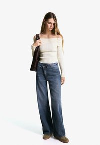 Off-the-shoulder cream ribbed knit top, blue wide-leg jeans, and brown suede shoes; model holding a dark brown leather bag.