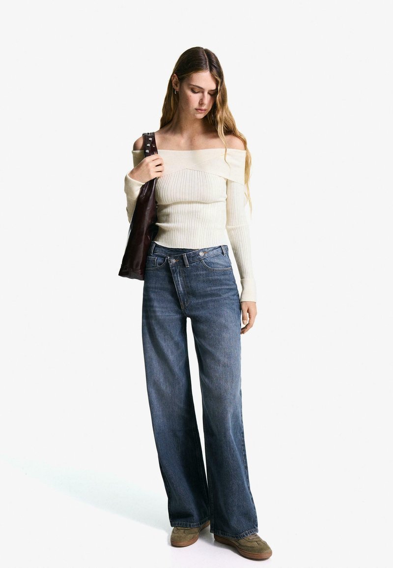 Off-the-shoulder cream ribbed knit top, blue wide-leg jeans, and brown suede shoes; model holding a dark brown leather bag.