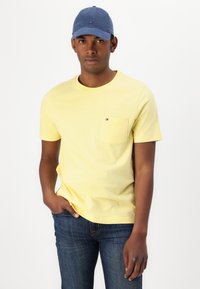 Gul bomulds t-shirt med rund hals og brystlomme, parret med mørkeblå denim jeans. Accessories inkluderer en blå baseball cap.