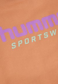 Gros plan sur un tissu orange avec un texte violet et bleu clair affichant "hummel SPORTSWEAR" en police moderne et en gras.