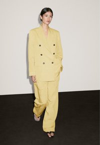 Ein Model läuft auf einem dunklen Boden und trägt einen gelben, doppelt geknöpften Anzug. Der Hintergrund ist hellgrau und hebt den Stil des Outfits hervor.