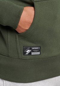 Olivgrön sweatshirt med ribbade muddarna och nederkanten. Har en framficka och en svart etikett med vit logotyp och text på den nedre vänstra sidan.
