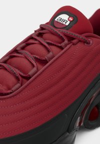 Red textile sneaker with ribbed design, featuring black accents and laces. Includes a circular logo on the tongue and a visible air unit.