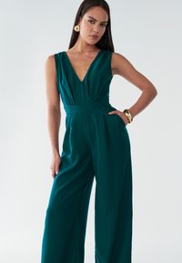 Teal jumpsuit med dyb V-udskæring, ærmeløs design, vidde ben og tætsiddende talje. Har glat stof og sidelommer. Gyldne accessories.