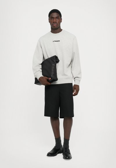 Lys grå hettegenser med svart logo, svart shorts, mørke grå sokker og svarte sko. Modellen holder en svart klut. Enkelt design.