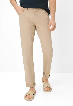 Mann trägt beige Chinos mit aufgekrempelten Saum und beige Sandalen mit gekreuzten Riemen, steht vor einem schlichten Hintergrund.