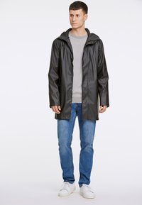 Svart vattentät regnjacka med huva, tillverkad av ett slätt material. Har tryckknappar och en lös passform, kombinerad med ljusblå jeans och vita sneakers.