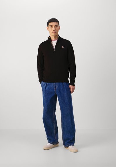 Fekete zipzáras pulóver, kisebb logóval, egy világos ing fölé rétegezve, laza kék farmernadrággal és bézs sportcipővel párosítva.