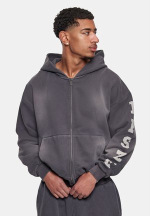 Noor mees, kellel on lühikesed lokkis juuksed, kannab tumedat halli lukuga dressipluusi, mille paremal varrukal on valged tähed, vaadates vasakule.