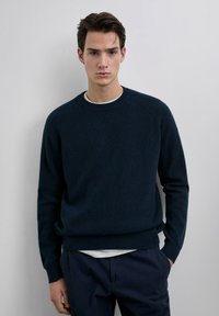 Dark navy sweater with ribbed texture, crew neckline, and long sleeves, paired with a white top and navy trousers. Simple and fitted design.