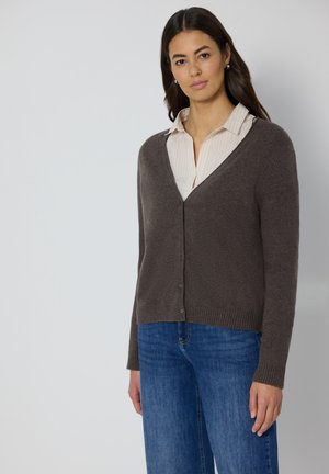 Brun cardigan med V-ringning, knappar fram och ribbad fåll, lager på lager över en ljus randig skjorta, kombinerad med blå jeans.