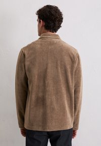 Bruine corduroy jas met een kraag, voorzien van een verticale ribbelstructuur en een rechte snit. Lijkt ontspannen met een achterste zoomlijn.