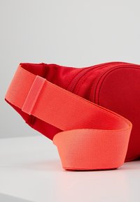 Červená látková ledvinka s texturovaným povrchem, vybavená jasně oranžovým nastavitelným popruhem a zipovou kapsou pro uložení.