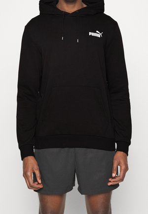 Person trägt einen schwarzen Puma Hoodie mit Fronttasche und Kordelzug, kombiniert mit dunkelgrauen Shorts, steht vor einem schlichten Hintergrund.