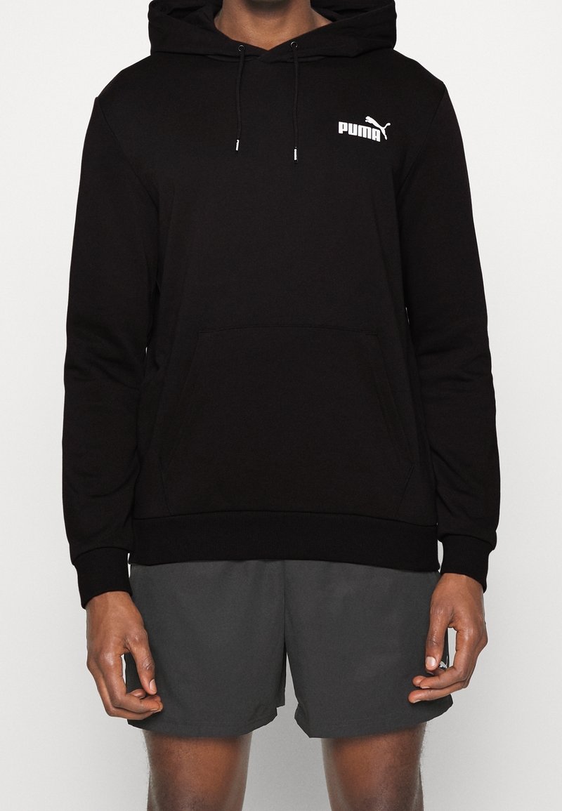 Personne portant un sweat à capuche noir Puma avec poche avant et cordons, associé à un short gris foncé, debout devant un fond uni.