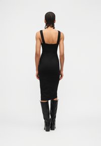 Γυναίκα με πλεγμένα μαλλιά, ντυμένη με εφαρμοστό μαύρο midi φόρεμα χωρίς μανίκια και μαύρες μπότες μέχρι το γόνατο, γυρισμένη προς τα πίσω σε απλό φόντο.