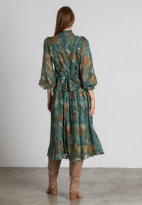 Vestido largo de color verde azulado, confeccionado con una tela ligera, que presenta patrones florales dorados, mangas abullonadas y un detalle de cintura atada. Longitud a mitad de pantorrilla.