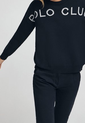 Personne portant un t-shirt noir à manches longues avec le texte "POLO CLUB" sur la poitrine et un pantalon noir sur un fond clair uni.
