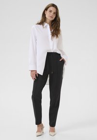 Ženska, ki modeli belo oversize srajco z gumbi, črne hlače na vrvico in bež visoke natikače, stoji na enobarvni svetli podlagi.