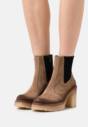 Hnědé semišové kotníkové boty s černým elastickým bočním panelem a robustním béžovým podpatkem. Mají zaoblenou špičku a texturovanou podrážku.