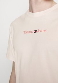 Camiseta de algodón de color rosa claro con cuello redondo, que presenta el bordado "Tommy Jeans" en letras rosas y negras en el pecho. Mangas cortas.