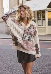 Kvinde med krøllet blond hår iført en mønstret strikket sweater og sort lædernederdel, står på en bygade med hånden berørende sit hår.