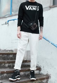 Persona che indossa un felpa nera Vans, pantaloni bianchi, sneakers alte in bianco e nero, borsa a tracolla con stampa leopardata, in piedi su scale all'aperto.