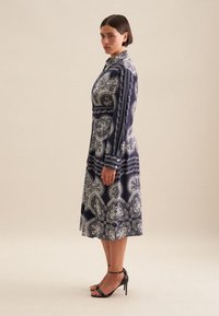 Abito navy con motivo paisley bianco, maniche lunghe, dettagli con bottoni e vita aderente. Abbinato a sandali neri con tacco alto.