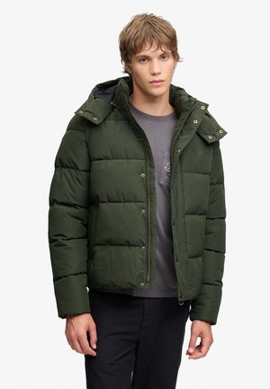 Young man wearing a dark green puffer jacket over a gray t-shirt and black pants, standing against a plain white background.