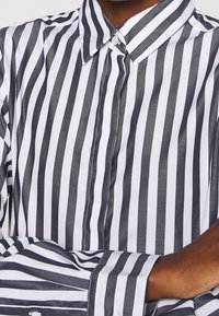 Svartvit randig skjorta med spetsig krage, knappstängning framtill och långa ärmar. Materialet verkar slätt och lättviktigt.