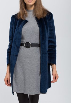 Manteau en laine bleu marine à coupe droite, avec des manches longues et un col fermé. Porté sur une robe grise, maintenue par une ceinture noire avec une boucle circulaire.