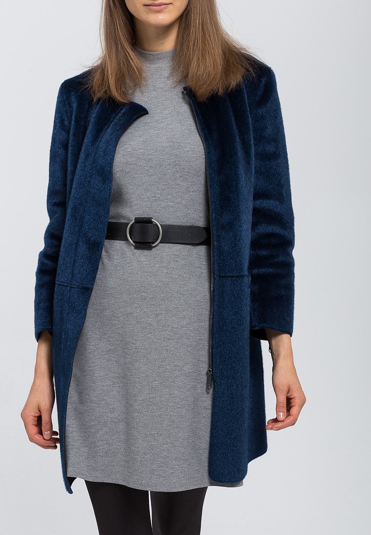 Mørkeblå ullkåpe med rett snitt, lange ermer og lukket krage. Båret over en grå kjole, festet med et svart belte med en rund spenne.