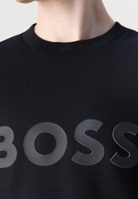 T-shirt nera con collo rotondo, caratterizzata da grandi lettere bianche lucide che spellano "BOSS" sul davanti; texture in cotone liscia.