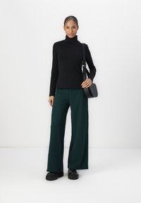 Zwarte langarm turtleneck trui, gecombineerd met groene broek met brede pijpen en chunky zwarte platform schoenen. Bijpassende zwarte schoudertas.