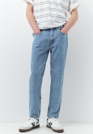 Oseba, oblečena v svetlo modre kavbojke z ravnimi hlačnicami, bele superge s črnimi detajli in belo majico z dolgimi črtami, z rokami v žepih.