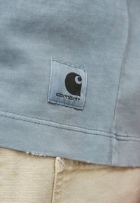 Jasnoniebieska koszulka z tkaniny z czarną łatą z logo Carhartt, naszywką oraz delikatnym teksturowanym detalem przy dolnym zakończeniu.