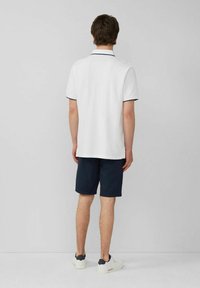 Weißes Poloshirt mit marineblauen Akzenten, kombiniert mit dunkelblauen Shorts. Modell von hinten gezeigt, trägt lässige weiße Sneakers. Flache, glatte Textur.
