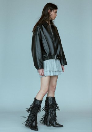 Jeune femme portant une veste en cuir noire oversize, une mini-jupe grise à volants et des bottes noires à franges montantes jusqu'aux genoux, debout de profil.