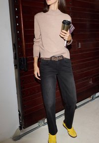 Mujer vestida con suéter beige, jeans negros con cinturón de leopardo y zapatillas amarillas, sosteniendo una taza de viaje y una revista frente a una puerta de madera.