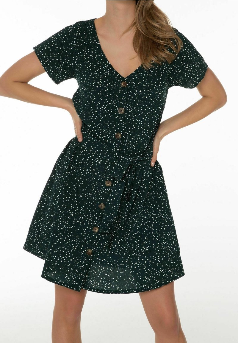 Robe vert foncé à pois blancs, manches courtes, col en V, et fermeture par boutons sur le devant. Comprend un détail de taille nouée.