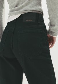 Jean en denim noir avec une coupe droite, comprenant deux poches arrière et une étiquette en cuir. Le tissu a une texture lisse.