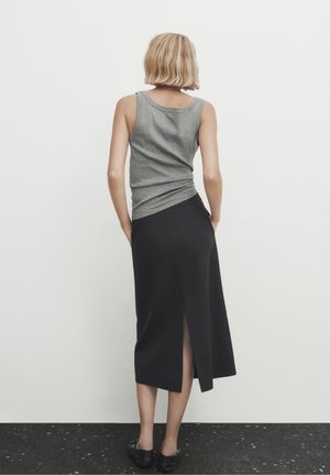 Vrouw met kort blond haar, gekleed in een grijs mouwloos topje en een zwarte midi-rok met een achterste split, staand tegen een effen witte muur.