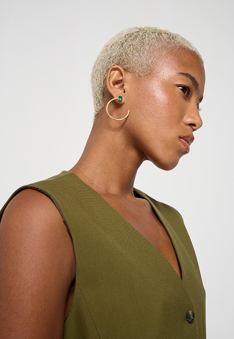 Mujer con cabello corto y rubio, que lleva una blusa sin mangas verde y un gran aro de oro con un pendiente de gema verde.