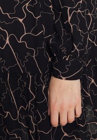 Schwarzes Kleid mit fließendem Design, das ein strukturiertes Muster aus hellrosa gewundenen Linien aufweist; beinhaltet geraffte Manschetten und einen lässigen Schnitt.